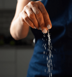 A person wearing a navy apron sprinkling some salt