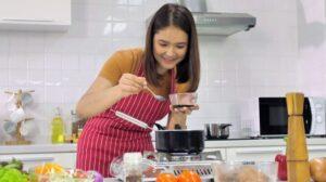 A woman putting in a teaspoon of sauce into a pot in the kitchen