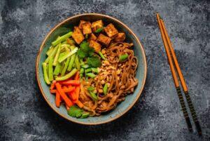 A bowl of noodles with fried tofu, cucumbers, carrots, and garnished with cilantro and spring onions