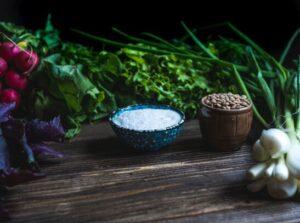 A small bowl of salt on a table, surrounded by different types of vegetables